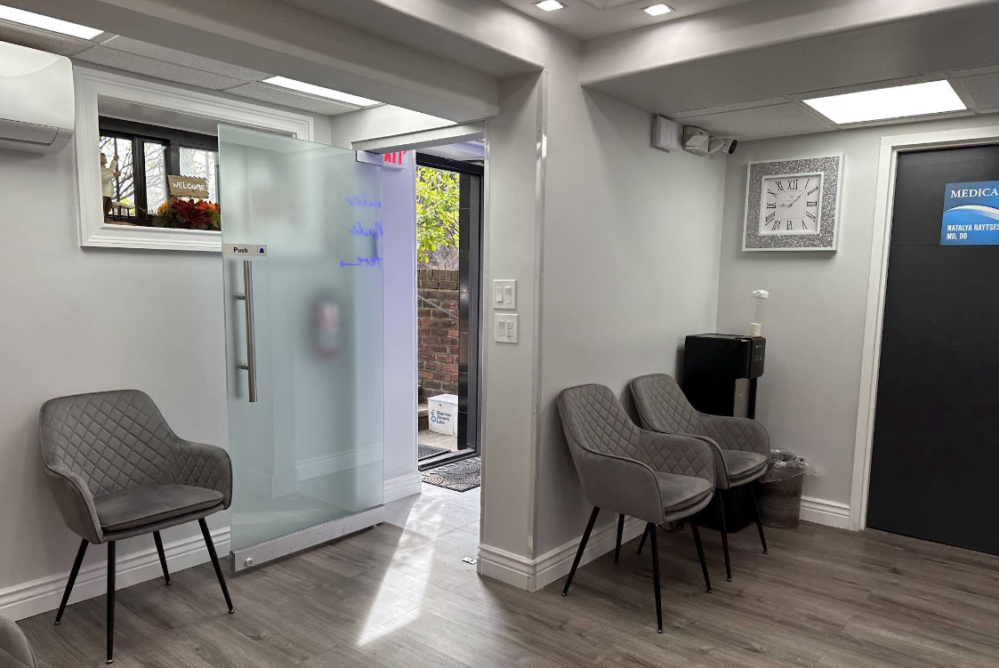 The image shows a modern dental office interior with a waiting area featuring two chairs, a glass partition, a reception desk with a computer monitor, and a window with blinds partially drawn.
