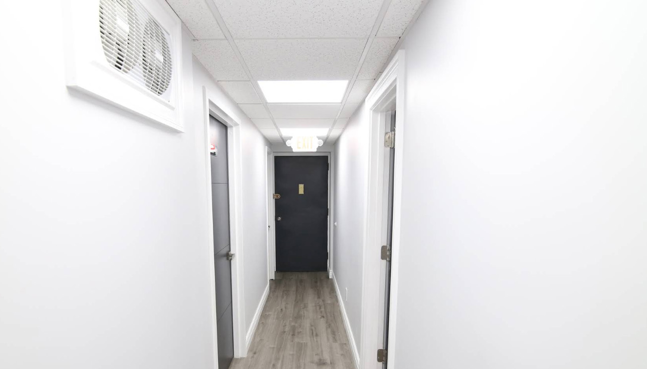 The image shows a long hallway with white walls and a light wood floor, featuring multiple doors on either side, leading to various rooms.