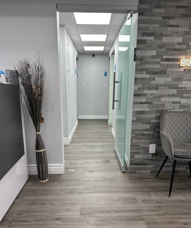 The image shows an interior hallway with a door on the right leading to a room with a glass wall. There is a vase with branches on a table, a white wall, and a modern decor style.