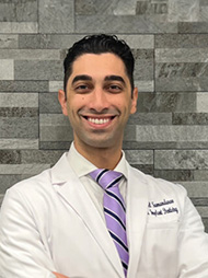 The image shows a man standing against a wall with a smile on his face, wearing a white lab coat with name tags, a purple tie, and a blue lanyard around his neck. He has a beard and mustache, and appears confident.