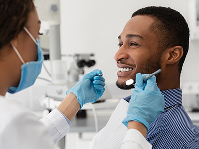 The image depicts a dental professional performing a dental procedure on a patient while wearing protective gloves and a surgical mask, with the patient smiling and looking towards the camera.