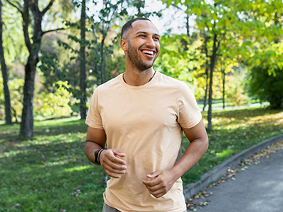 A man is jogging in a park with trees and grass around him, smiling and looking towards the camera.