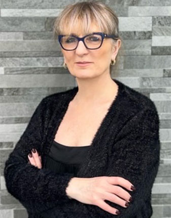 The image shows a woman standing against a gray wall with a brick pattern. She is wearing glasses, has short hair, and is dressed in a black top with her arms crossed.