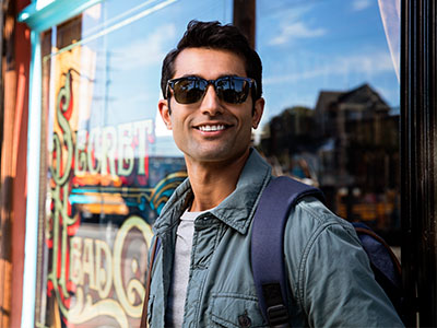 A young man wearing sunglasses, a backpack, and a casual jacket stands confidently outdoors with a smile, posing for a photograph by a storefront window.