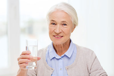 The image shows a woman holding a glass of water with both hands, smiling slightly, and looking directly at the camera.