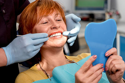 A woman with red hair sitting in a dentist s chair, smiling while holding a blue mouthguard, with a dental professional assisting her by placing a stethoscope on her chest.