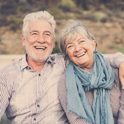 The image shows a woman with a radiant smile, leaning on her arm with her elbow resting on a wooden surface, against a neutral background.