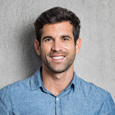The image shows a smiling man with short hair, wearing a blue denim shirt, standing against a grey wall background.