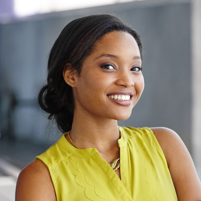 The image shows a woman smiling at the camera with her hair pulled back, wearing a yellow top and a necklace, standing against a blurred background that appears to be an indoor setting.