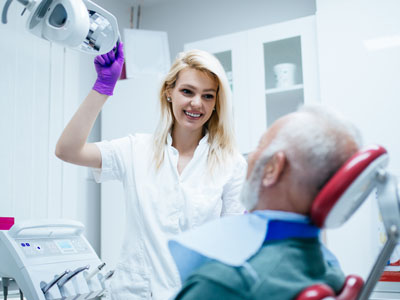 A dental hygienist in a white uniform adjusting a patient s chair while the patient sits with their eyes closed, wearing a surgical face mask.
