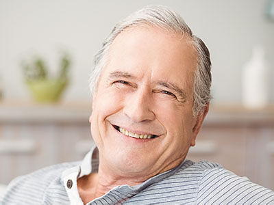 The image shows a smiling older man with gray hair, wearing glasses and a blue shirt, sitting comfortably with his arms crossed.