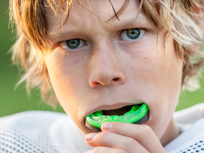 A young male athlete with blonde hair, wearing a football uniform, holding a green sports mouthguard close to his mouth.