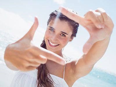 The image shows a woman with long hair smiling at the camera while holding up a finger to frame her face. She appears to be outdoors, possibly on a beach, given the bright daylight and the ocean in the background.